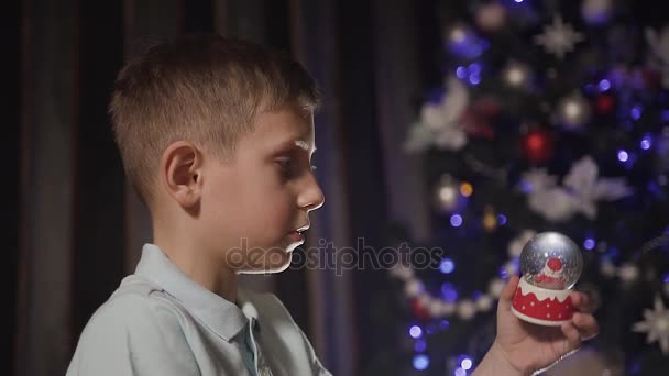 Close-up. Op een kerstfeest speelt een kleine jongen met een sneeuwbol binnen die santa is — Stockvideo