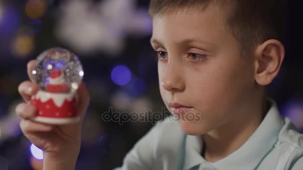 Närbild pojken håller i hans händer en snowjordklot, i mitten av som det är en tomte, och astonishedly skakar den. Julgran, Leksak, holiday — Stockvideo