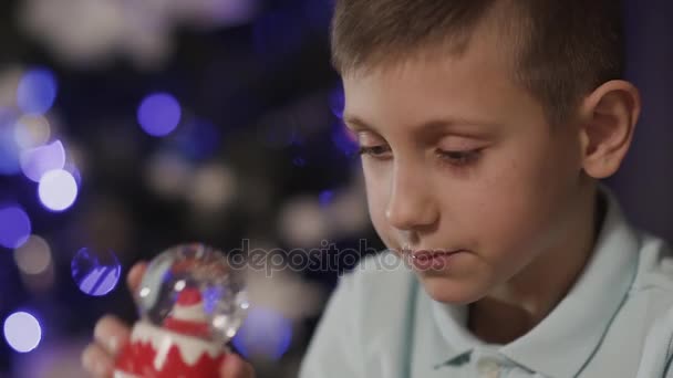 Close-up op de achtergrond van een wazig kerstboom, schudt de jongen een decoratief speelgoed bal die in het midden van die de Kerstman is — Stockvideo