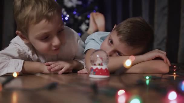 Primer plano de dos niños tumbados en el suelo cerca del árbol de Navidad y sacudiendo una bola de nieve en medio de la cual está el Santa Claus. Concepto de regalos de invierno y Navidad — Vídeos de Stock