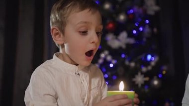 Plan - arka plan bulanık bir Noel ağacı üzerinde oturma odasında katta oturan ve darbeler uzak bir iltihaplı yeşil mum tutan yakışıklı bir küçük çocuk kapatın