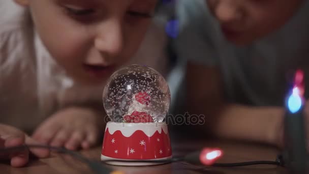 Close-up Dua anak mengguncang bola salju, meletakkannya di lantai dan mengintip di dalamnya saat salju turun di santa claus terletak di dalam bola salju ini — Stok Video