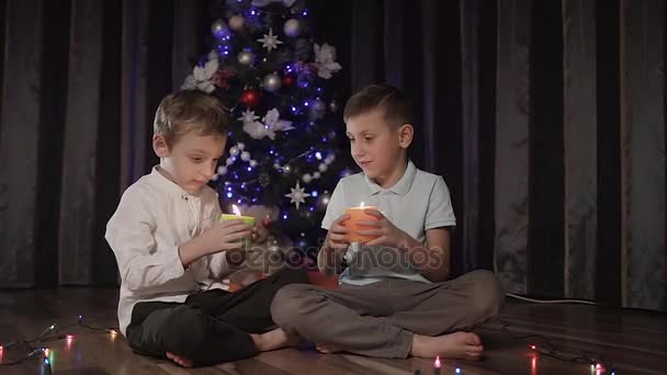 Dois caras caucasianos estão sentados na sala no chão perto de árvores de Natal e segurando velas decorativas verdes e laranja nas mãos — Vídeo de Stock