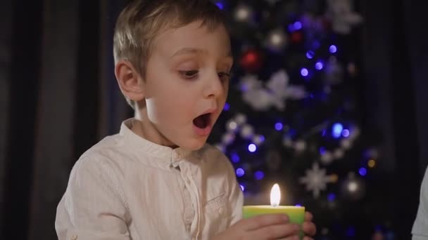 Tutup rencana - di latar belakang pohon Natal kabur seorang anak kecil yang tampan duduk di lantai di ruang tamu dan memegang lilin hijau yang membara yang meniup jauh — Stok Video