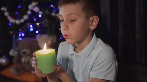 Close plan - auf dem Hintergrund eines verschwommenen Weihnachtsbaums sitzt ein hübscher kleiner Junge auf dem Boden im Wohnzimmer und hält eine entzündete orangefarbene Kerze in der Hand, die wegweht — Stockvideo