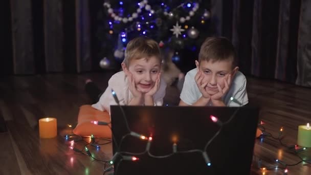 Meninos bonitos relaxando com seu laptop na frente de uma árvore de Natal deitada descalça no chão cercada por guirlandas coloridas e velas que brilham à noite — Vídeo de Stock