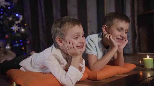 Plano do meio - no fundo de uma árvore de Natal azulada, dois irmãos estão no quarto no chão sobre os travesseiros laranja e assistir a um desenho animado engraçado no laptop — Vídeo de Stock