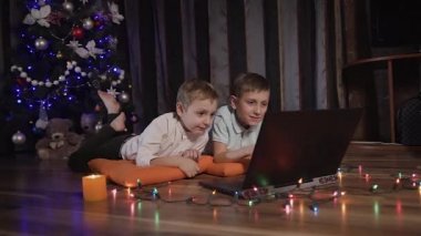 Yeni yıl akşam oturma odasında iki çocuk üzerinde dekoratif yastıklar yalan ve bir dizüstü bilgisayarda komik bir çizgi film izlemek. Renkli çelenk yere koyulur