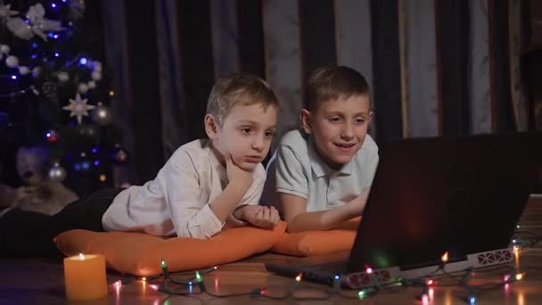 Dois lindos garotinhos estão deitados em almofadas decorativas laranja no chão na sala de estar e assistindo a um desenho animado em um laptop através de uma rede social. No chão na frente das crianças deitado — Vídeo de Stock