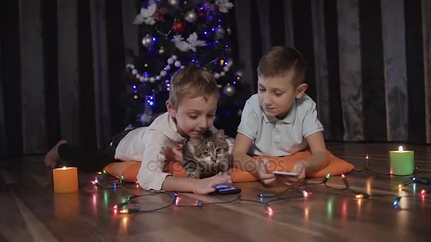 Dois meninos e seu gato estão deitados no quarto no chão antes da árvore de Natal com seu pequeno gato e jogando jogos em seus telefones pela internet — Vídeo de Stock