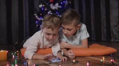 Yakın plan - iki küçük kardeş süslü turuncu yastıklar Noel ağacı yakınındaki oturma odasında yere yalan ve onların gadget'ı çevrimiçi oyunları oynayabilirsiniz. Renkli çelenk