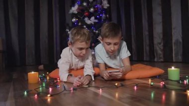 Arka planda bir Noel ağacı, iki çocuk yerde portakal yastıklar üzerinde yalan ve sosyal ağ üzerinden telefonda oyna