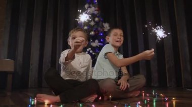 Bir Noel ağacı yakınındaki bir katta oturan ve yanan bir köpüklü şişe tutan ve mutlu bir şekilde gülümseyen güzel çocuklar