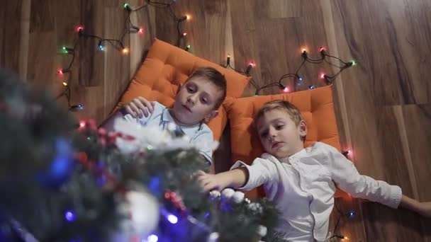 Twee kleine jongens hebben plezier op oranje kussens in de buurt van een decoratieve kerstboom liggen. Gekleurde slingers verspreid op de vloer in de buurt van de kinderen — Stockvideo