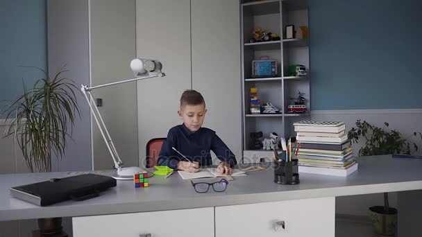 Kleiner süßer Junge in blauem Hemd, der an einem Schreibtisch in seinem Zimmer sitzt und Hausaufgaben macht. Bücher auf dem Tisch, Hausaufgaben — Stockvideo