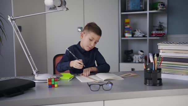 Un chico colegial está vestido con una camisa azul oscuro sentado en la mesa en casa y haciendo una tarea. Libros y cuadernos están sobre la mesa — Vídeos de Stock