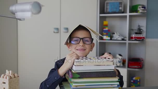 Pequeno aluno de óculos tem muitos livros, colocar um caderno em sua cabeça e shows divertidos com uma mão que está tudo bem. TPC. Estudante alegre em casa — Vídeo de Stock