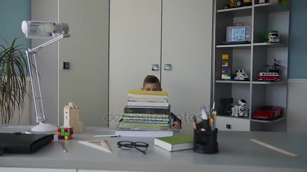Primer plano de un guapo chico divertido que, corre al escritorio detrás del cual hace los deberes y abraza sus muchos libros con diversión. Tareas — Vídeo de stock