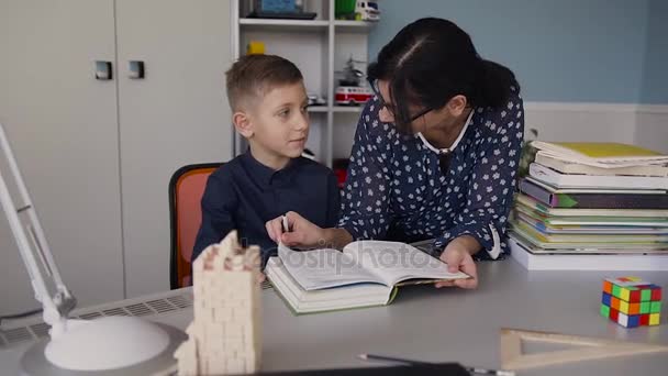 Un chico guapo con una camisa azul oscuro lee un libro interesante con su profesor usando gafas. Un estudiante realiza la tarea en casa — Vídeos de Stock