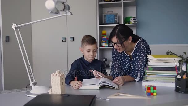 Lesen mit der Lehrerin - ein Grundschüler liest ihrer Lehrerin vor. Lehrer sitzt am Schreibtisch neben männlichen Studenten, während sie zusammen lesen. Schüler und Lehrer lesen gemeinsam ein Buch im weißen Raum — Stockvideo