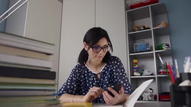 A estudante de óculos sentada na mesa segura o celular em casa. Menina bonita sentada à mesa com livros usando telefone celular e sorrindo — Vídeo de Stock