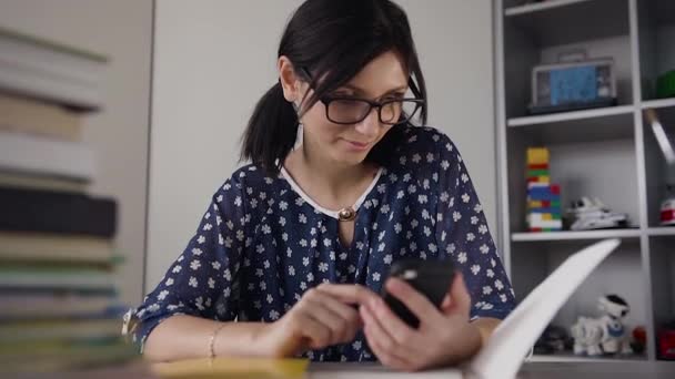 Школярка в окулярах, сидячи за столом, тримає мобільний телефон вдома. Красива дівчина сидить за столом з книгами, використовуючи мобільний телефон і посміхаючись — стокове відео