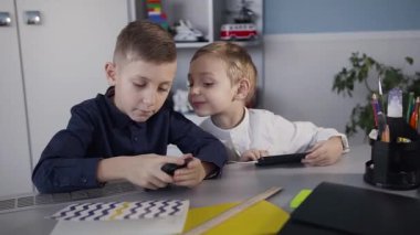Küçük çocuklar evde beyaz oda, masada oturan ve iskambil spor üstünde onların telefon. Beyaz bir gömlek küçük bir çocuk telefonda online oyna eğleniyor