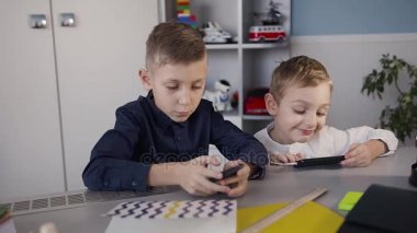 Küçük çocuklar evde beyaz oda, masada oturan ve iskambil spor üstünde onların telefon. Beyaz bir gömlek küçük bir çocuk telefonda online oyna eğleniyor
