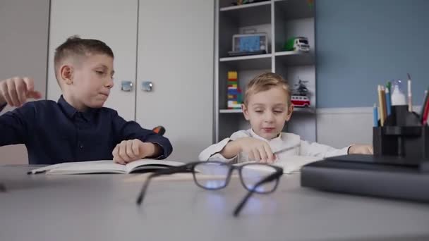 Faceci nie odrabianiu i radośnie dostanie siebie nawzajem w swoich dłoniach. Dzieci siedzą przy biurku w domu w białym pokoju. Szkolne lekcje w domu robi — Wideo stockowe