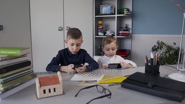 Dos chicos están sentados en la mesa en la habitación blanca y juegan un juego en línea en sus teléfonos. Niño caucásico joven jugando juegos en línea en el teléfono inteligente — Vídeos de Stock