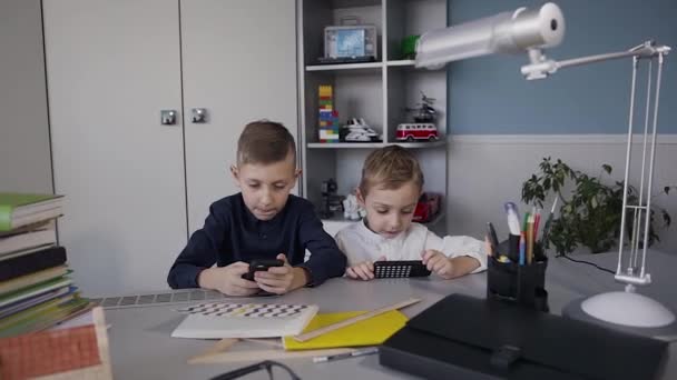 Bei bambini dipendenti dai telefoni cellulari che giocano con facce concentrate. Due ragazzi che usano il telefono per giocare online a casa seduti al tavolo — Video Stock