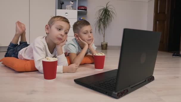 オレンジ色の装飾的な枕に床の上に横たわる美しい男の子、おいしいポップコーンを食べるし、ノート パソコンで無料インターネットのネットワークを通して面白い漫画を見る — ストック動画