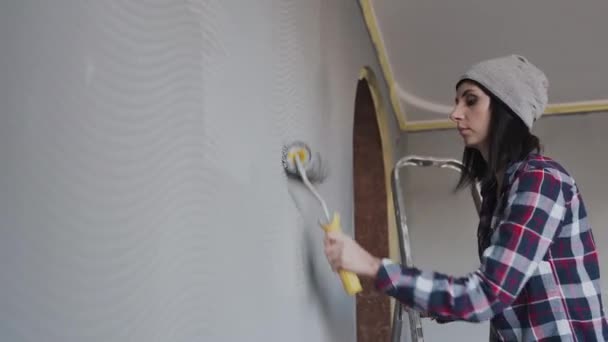 En ung hipsters flicka klädd i en grå hatt, står på stegen i rummet och målar väggarna i sovrummet i en grå färg. Kvinnan håller en rulle i en grå färg — Stockvideo