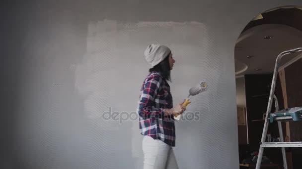Uma jovem mulher em um chapéu cinza desenha uma parede em sua nova casa na cor cinza, ouve música do telefone em fones de ouvido e dança divertida — Vídeo de Stock