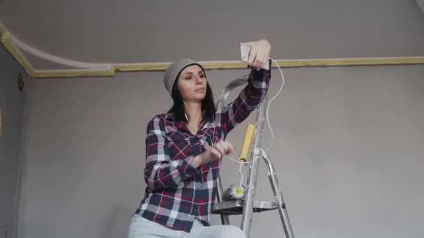 Une jeune fille est assise sur l'échelle et fait un selfie dans la pièce où elle peint les murs d'une couleur grise. Réparation dans l'appartement — Video