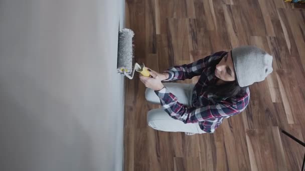 Ovanifrån - en ung vacker flicka i grå hatt drar väggarna med en roller i grå färg. En kvinna gör reparationer i sin nya lägenhet — Stockvideo