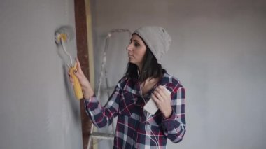 Gri şapkalı hippi kız boyama duvar rulo gri renkte çizmek ve telefondan kulaklık müzik dinler. Kapalı onarım