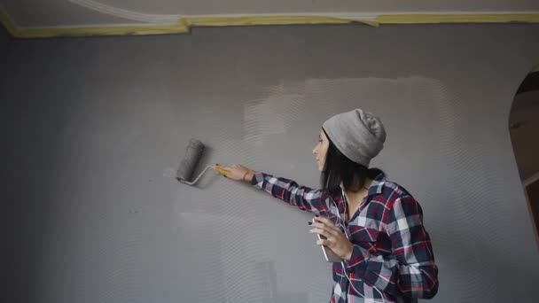 Una joven con un sombrero gris dibuja una pared en su nuevo hogar a color gris, escucha música desde el teléfono con auriculares y baile divertido — Vídeos de Stock