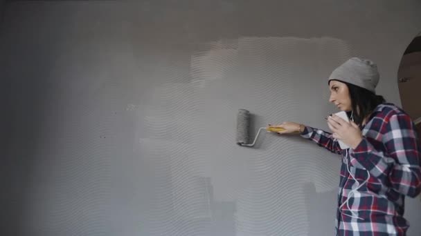 Krásná mladá dívka poslouchá hudbu z telefonu sluchátka a dokončí, malování součástí Velké zdi s šedou barvou a Kartáčový válec. Žena omítkou zdi doma — Stock video