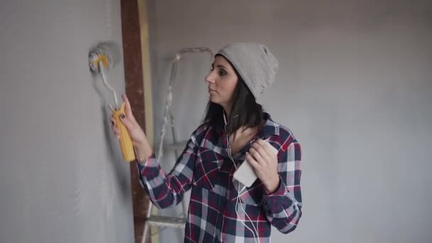 La chica hipster en sombrero gris dibuja la pared de pintura con rodillo en gris y escucha música en los auriculares desde el teléfono. Reparación interior — Vídeo de stock