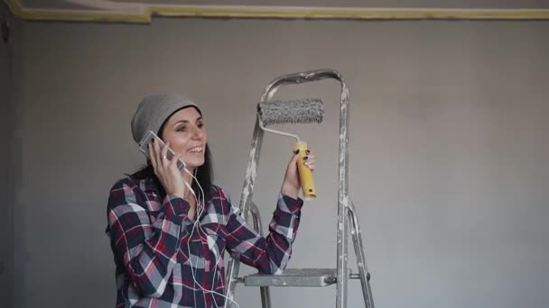 年轻, 时髦的女孩坐在梯子上的房间里, 她用灰色的颜色绘制墙壁, 并在电话中说有趣的。在卧室里修理。粉刷房间的墙壁 — 图库视频影像
