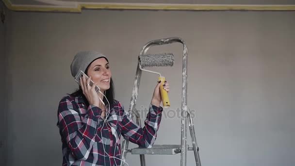 Junges Hipstermädchen, das auf der Leiter in dem Raum sitzt, in dem sie die Wände in grauer Farbe bemalt und Spaß am Telefon hat. Reparatur im Schlafzimmer. Tünche die Wände im Raum — Stockvideo