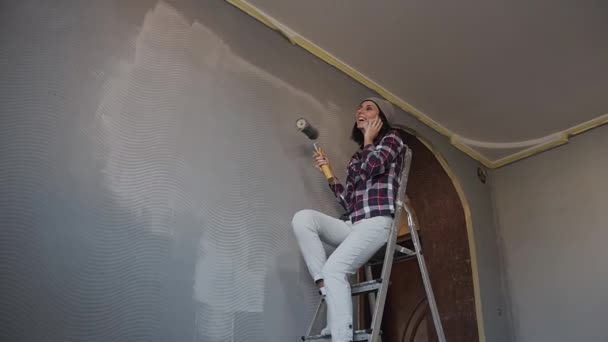 Jeune fille hipster assise sur l'échelle dans la pièce où elle peint les murs d'une couleur grise et parle amusant au téléphone. Réparation dans la chambre. Blanchir les murs dans la chambre — Video