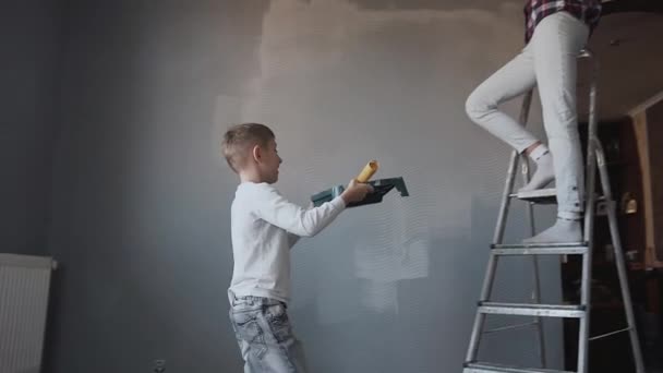 Een jonge vrouw staande op een ladder in de buurt van de muur, geeft haar zoon haar een container met een grijze verf en een roller zodat ze van de kleur van de muur veranderen kan. Reparatie in het huis. Witten de — Stockvideo