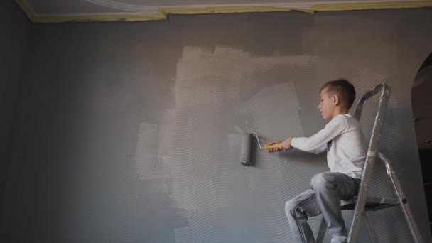 Ребенок садится на лестницу в комнате и рисует стену с помощью ролика серого цвета. Парень отбеливает стену в новой комнате. Счастливый мальчик — стоковое видео
