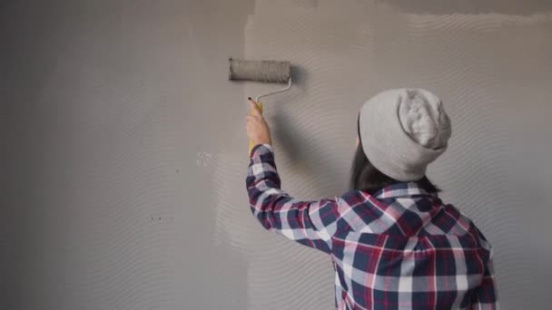 Bokovky mladá dívka oblečená v šedém klobouku, stojí na žebříku v místnosti a maluje zdi v ložnici v šedé barvě. Žena drží váleček v šedé barvě — Stock video