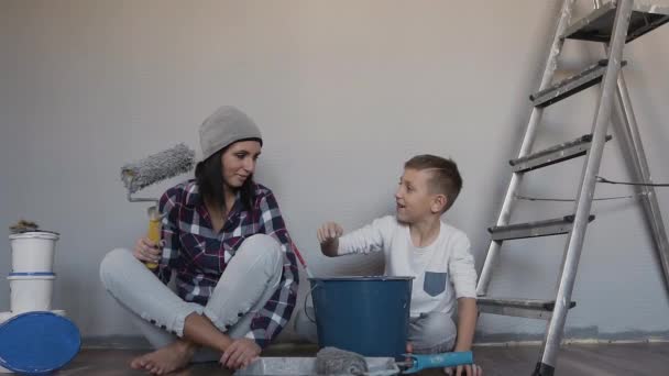 Mãe e filho terminaram de reparar no quarto eles se sentam no chão e pintam uns aos outros com tinta. Uma mulher segura um rolo na mão. Feliz dia acabado de reparar — Vídeo de Stock