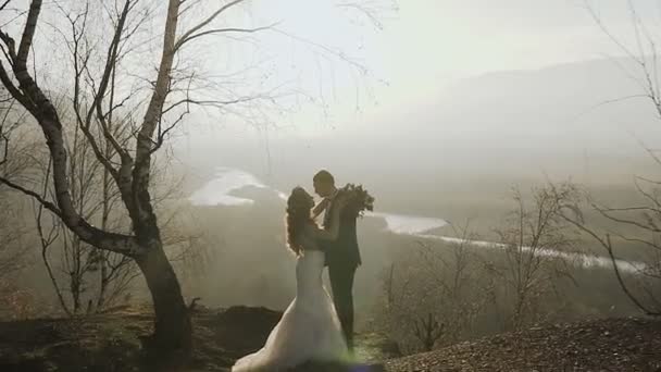 A noiva e o noivo estão no alto das montanhas ao pôr-do-sol no fundo do rio. Concepção outono ou primavera — Vídeo de Stock