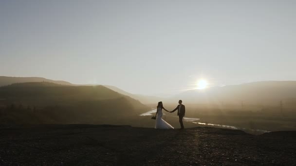 Puesta de sol en las montañas. Hermosa novia joven y prometido camina en la puesta de sol en el fondo del río y el bosque. Concepción del día de la boda. Noche romántica — Vídeo de stock