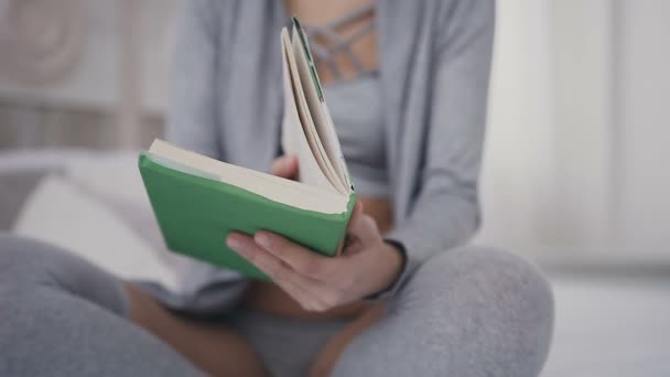 Красивая брюнетка в очках сидит на кровати и держит книгу для чтения. Молодая женщина открывает интересную книгу в зеленой обложке дома — стоковое видео